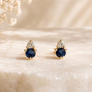 May include: A pair of stud earrings crafted in gold, each featuring a round, deep blue sapphire stone at the base topped with a smaller, clear, round-cut diamond. The stones are held in place by gold prongs, creating a vertical, two-stone design.
