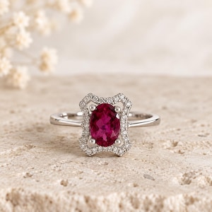 Peut inclure: Une bague en argent avec une pierre précieuse ovale rose foncé au centre, maintenue par quatre griffes. La pierre est entourée d'un cadre rectangulaire décoratif serti de petits diamants pavés blancs, sur un anneau en argent poli.