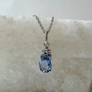Op de afbeelding: Een zilverkleurige hanger aan een ketting met een lichtblauwe edelsteen in ovale slijpvorm, geplaatst in een metalen zetting met vier pootjes. De hanger is aan de bovenzijde versierd met twee kleine heldere steentjes en hangt aan een fijne ketting.
