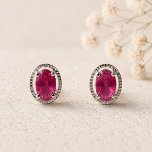 May include: A pair of stud earrings featuring oval-cut vibrant pink gemstones set in silver-toned metal. Each oval stone is surrounded by a textured, grooved halo frame and secured with four prongs, creating a classic and symmetrical jewelry design.