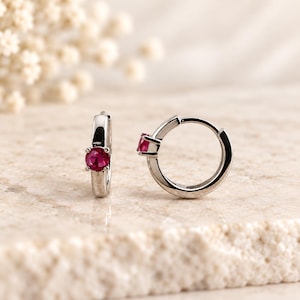 Può includere: Un paio di orecchini a cerchio color argento, ciascuno caratterizzato da una singola pietra preziosa rotonda di un vivido colore rosso rosato, montata su una struttura a quattro griffe. I cerchi hanno una finitura metallica lucida.