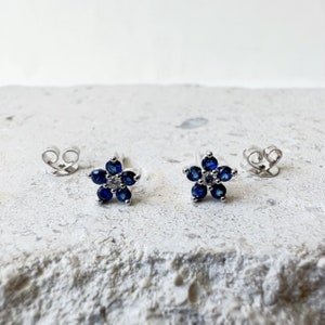 Puede incluir: Un par de pendientes de bot&oacute;n de plata con un dise&ntilde;o de flor. Cada flor est&aacute; hecha de piedras azules y una sola piedra blanca en el centro.