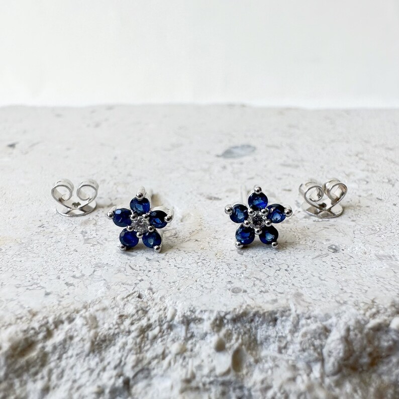 Puede incluir: Un par de pendientes de bot&oacute;n de plata con un dise&ntilde;o de flor. Cada flor est&aacute; hecha de piedras azules y una sola piedra blanca en el centro.