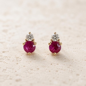 May include: A pair of stud earrings featuring a round, faceted deep pink ruby gemstone set in gold, topped with a smaller, round, clear white diamond. Each earring is secured with a gold setting that holds both stones in a vertical, tiered arrangement.