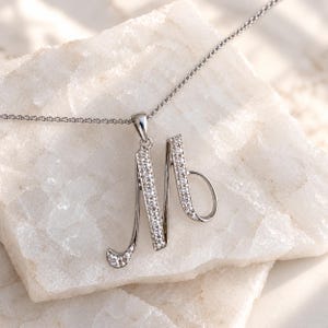 May include: A silver-toned necklace featuring a cursive letter M pendant. The pendant is embellished with numerous small, clear, sparkling stones set along the front surface of the letter. The chain is a delicate, thin, silver-toned metal link style.