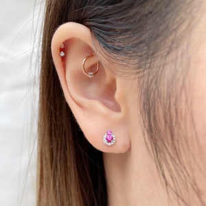 May include: A close-up of a person's ear with a pink gemstone earring with a halo of smaller white gemstones. The earring is in the lower lobe of the ear.