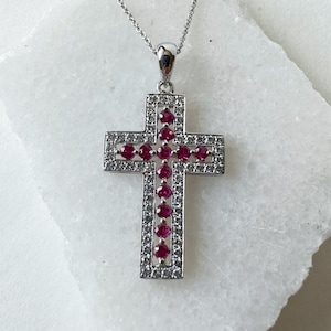 Puede incluir: Un colgante de cruz de plata con un diseño de piedras blancas y rojas. La cruz está adornada con pequeñas piedras redondas rojas engastadas en un marco de plata. El colgante está rodeado por un halo de piedras blancas más pequeñas.