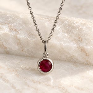 Può includere: Una collana color argento con un ciondolo a forma di rubino rotondo incastonato a lunetta. Il ciondolo è sospeso a una delicata catena a maglie, mostrando una gemma circolare rosso vivo incastonata in una montatura metallica liscia e lucida.