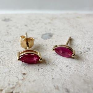 Puede incluir: Un par de pendientes de oro con gemas rojas oscuras en forma de marquesa. Los pendientes tienen un diseño simple y elegante con un marco dorado alrededor de cada gema. Los pendientes están colocados sobre un fondo neutro y texturizado.