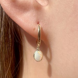 May include: Gold hoop earrings with a dangling opal gemstone. The opal is white with a hint of iridescent color.
