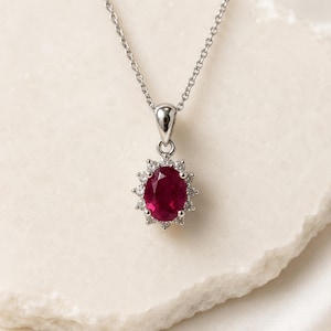 May include: A silver-toned necklace featuring an oval-shaped, deep red gemstone pendant. The central red stone is surrounded by a halo of small, clear, sparkling stones set in a metallic frame, suspended from a delicate, thin silver-toned chain.