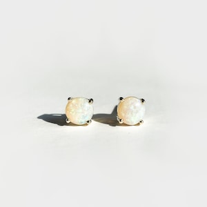 May include: A pair of opal stud earrings. Each earring features a round, iridescent opal stone set in a silver-colored metal. The opals display a mix of white, cream, and subtle flashes of color. The earrings are displayed on a white background.