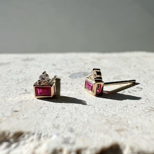 May include: A pair of gold stud earrings. Each earring features a rectangular, deep pink gemstone set in a gold frame, topped with a small, clear gemstone. The earrings are displayed on a textured, light-colored surface.