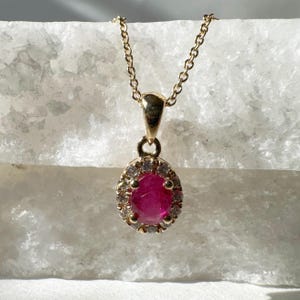 Puede incluir: Un collar de oro con un colgante que presenta una vibrante piedra preciosa roja rodeada de pequeñas piedras preciosas claras. El colgante tiene forma ovalada y cuelga de una delicada cadena de oro. El collar se muestra sobre un fondo blanco texturizado.