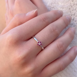 May include: A delicate silver ring with a small flower design featuring red and white gemstones.