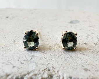 Puede incluir: Un par de pendientes de botón con piedras preciosas ovaladas de color verde oscuro. Las piedras están engastadas en un metal dorado con cuatro garras. Los pendientes se muestran sobre una superficie clara y texturizada.