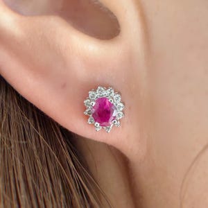 May include: A close-up of a ruby and diamond stud earring. The earring features a vibrant oval-cut ruby surrounded by a halo of small, sparkling diamonds. The setting appears to be white gold or platinum, creating a classic and elegant look.