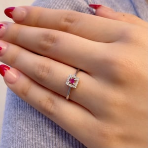 Pode incluir: Um anel de prata com uma cravação quadrada com uma gema vermelha vibrante no centro, rodeada por pedras brancas menores e brilhantes. O anel é exibido em uma mão, mostrando seu design delicado e apelo elegante.
