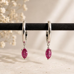 May include: A pair of silver hoop earrings featuring a polished metallic finish. Each hoop is adorned with a single marquise-cut, vibrant pink gemstone dangling from the bottom, secured by a small silver link that allows for subtle movement.