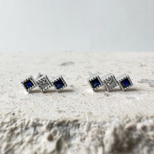 May include: A pair of silver earrings with three square-shaped blue and white gemstones. The gemstones are set in a silver frame with a textured edge.