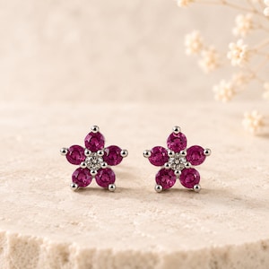 May include: A pair of stud earrings featuring a flower design. Each earring is crafted with five round, deep pink gemstones forming the petals and a single, smaller clear, sparkling gemstone set in the center, all held in a silver-toned metal.