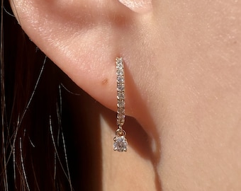 May include: A gold hoop earring with a single diamond dangling from the bottom. The earring is set with small diamonds around the hoop.