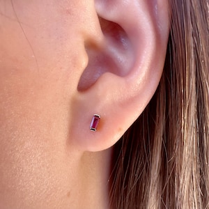 May include: A close-up of a person's ear with a small, rectangular, red gemstone earring.
