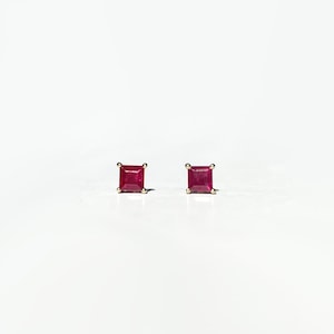 May include: A pair of square-cut ruby stud earrings. The vibrant red gemstones are set in gold, creating a classic and elegant look. The earrings are displayed against a clean, white background, highlighting their color and design.