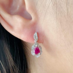 May include: A close-up of a dangling earring featuring a teardrop-shaped, deep red gemstone. The gemstone is encircled by a halo of small, clear stones. The earring has a floral design and is set in a silver-coloured metal.