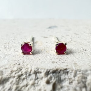 May include: A pair of gold stud earrings with small, round, red ruby gemstones.