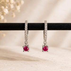 May include: A pair of silver-toned hoop earrings featuring a row of small clear stones along the front of the hoop and a single round, vibrant pink gemstone suspended from the base of each earring in a four-prong setting.