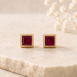 May include: A pair of stud earrings featuring square-cut deep red gemstones set in polished gold-toned metal frames. The earrings have a clean, geometric design with a smooth, reflective surface on the gold border surrounding the central stone.