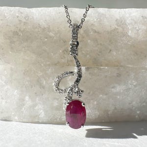 Puede incluir: Un collar de plata con un colgante de rubí y detalles de diamantes. El colgante tiene forma de una letra 'S' estilizada y cuelga de una cadena delicada.