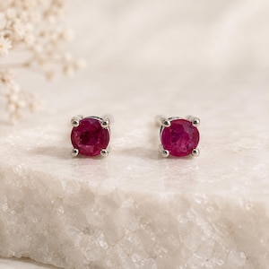 May include: A pair of round-cut ruby stud earrings set in silver-toned metal. Each earring features a vibrant, deep red gemstone secured by four prongs, showcasing a faceted surface that reflects light, presented as a matching set of two.