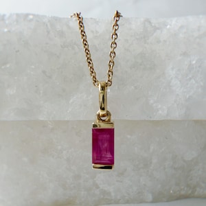 May include: A gold-colored necklace with a rectangular, ruby-colored pendant. The pendant is set in a gold frame and hangs from a delicate gold chain. The necklace is displayed against a white background.
