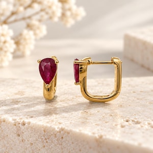 May include: A pair of gold hoop earrings featuring a pear-shaped, deep red ruby gemstone set at the front of each hoop. The earrings have a polished gold finish and a hinged closure mechanism, showcasing a vibrant red stone contrast.