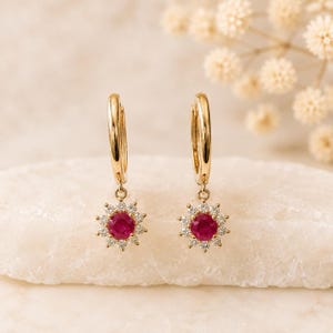 May include: A pair of gold hoop earrings featuring a dangling charm shaped like a flower. Each charm has a round, deep red ruby at the centre, surrounded by a halo of small, clear, sparkling white diamonds set in a gold metal frame.
