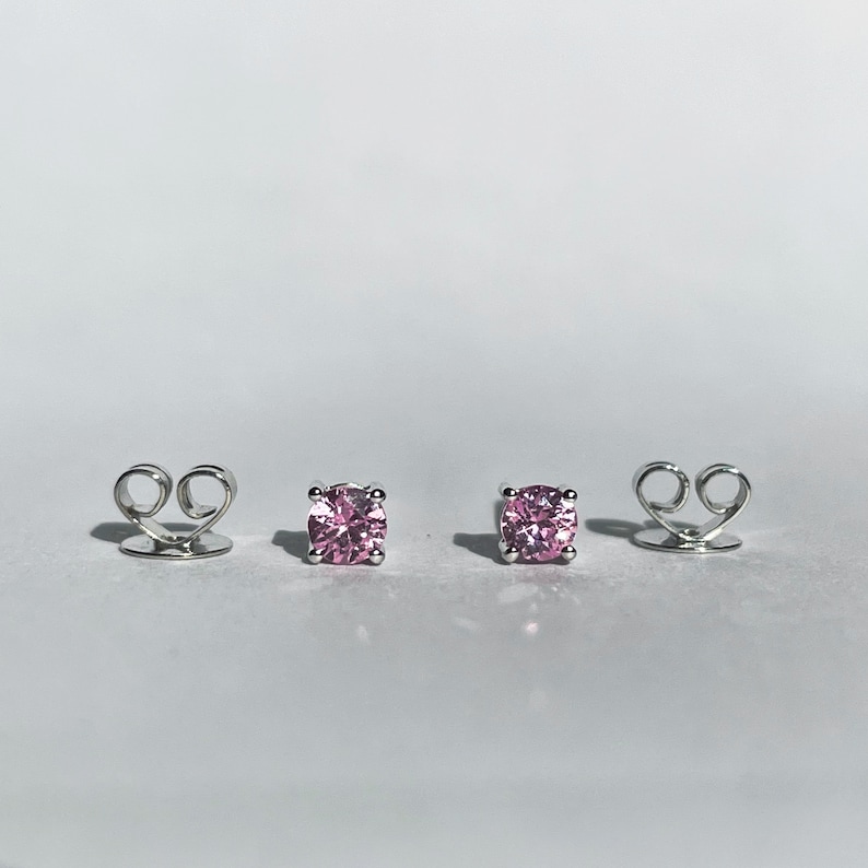 May include: A pair of stud earrings featuring round-cut pink stones set in four-prong silver-toned metal settings, accompanied by two matching silver-toned metal butterfly-style earring backs positioned separately on either side of the studs.
