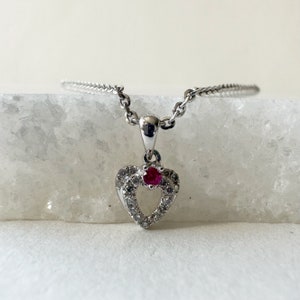 May include: A silver heart-shaped pendant necklace with a small red gemstone in the center and smaller clear gemstones around the edge.