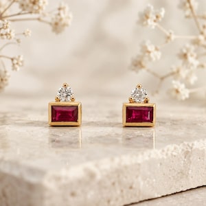 May include: A pair of gold stud earrings, each featuring a rectangular baguette-cut deep red ruby gemstone set horizontally, topped with a single round brilliant-cut white diamond in a prong setting, creating a refined and elegant jewelry piece.