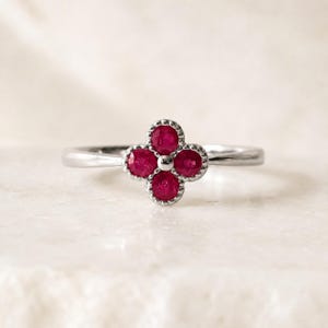 May include: A silver-toned ring featuring a four-petaled flower motif at the center. The flower is composed of four round, faceted red rubies set within a beaded metal frame, attached to a smooth, polished silver-toned metal band.