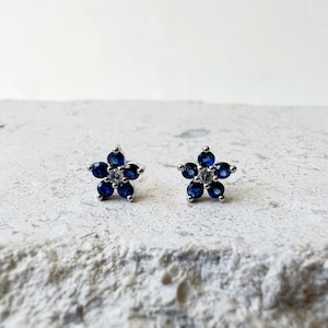Puede incluir: Un par de pendientes de bot&oacute;n de plata con un dise&ntilde;o de flor. La flor est&aacute; hecha de zafiros azules y un solo diamante blanco en el centro.