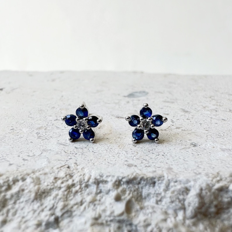 Puede incluir: Un par de pendientes de bot&oacute;n de plata con un dise&ntilde;o de flor. La flor est&aacute; hecha de zafiros azules y un solo diamante blanco en el centro.