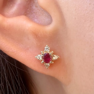 Puede incluir: Un pendiente de oro con una gema roja oscura redonda en el centro, rodeada de gemas claras más pequeñas. El pendiente tiene un diseño de estrella, con las gemas engastadas en un engaste de metal dorado. Se muestra en una oreja.