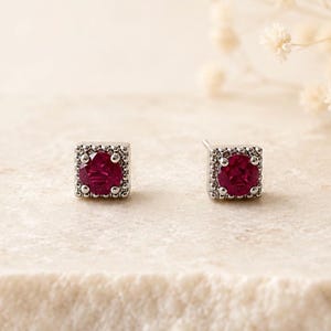 May include: A pair of stud earrings featuring a round, deep red gemstone set within a square, silver-toned metal frame. The square border is decorated with a series of small, raised metallic beads, creating a textured edge around the central stone.