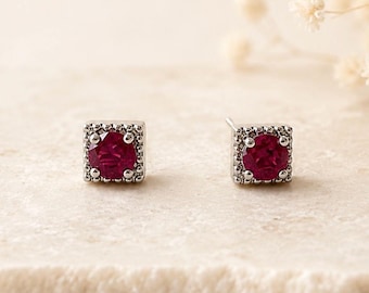 May include: A pair of stud earrings featuring a round, deep red gemstone set within a square, silver-toned metal frame. The square border is decorated with a series of small, raised metallic beads, creating a textured edge around the central stone.