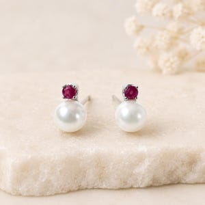 May include: A pair of stud earrings featuring a round white pearl base topped with a small, round, faceted deep red ruby gemstone set in a silver-toned metal prong setting. The earrings are designed with a post back for secure attachment.