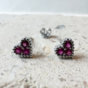 May include: A pair of heart-shaped stud earrings with three magenta-colored gemstones set in a silver-colored metal. The earrings are displayed with a single earring back.