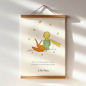 May include: A framed print of an illustration from "Le Petit Prince." The image shows a boy with yellow hair and a yellow scarf, sitting with an orange fox. The print includes French text and is held by a wooden frame.