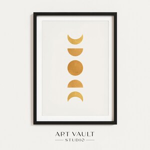 Pode incluir: Impressão de arte emoldurada com um design de fases da lua douradas sobre um fundo branco. As fases incluem formas de crescente, meia lua e lua cheia. A impressão está em uma moldura preta, com o texto "ART VAULT STUDIO" na parte inferior.